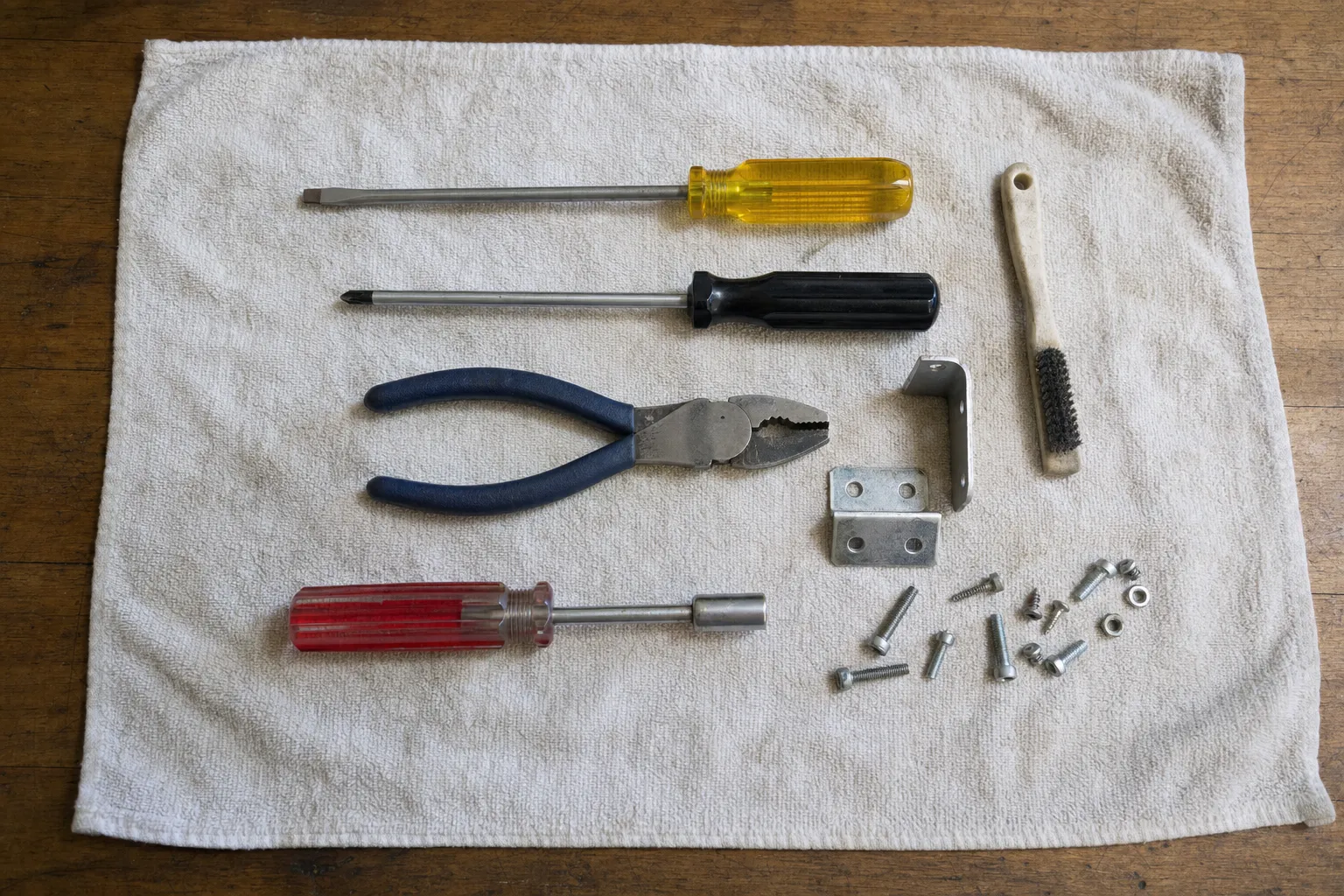 Screwdrivers, pliers, a nut driver and a wire brush laid out with mounting brackets, screws and nuts on a towel.